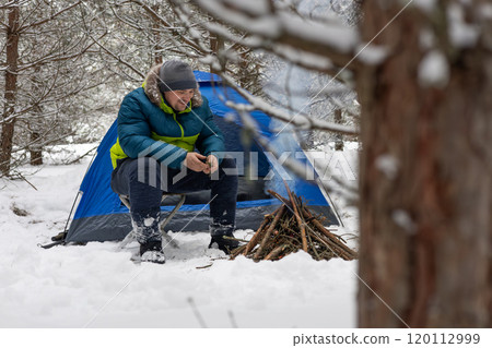 A camper man collects firewood for a fire near his tent in a snowy forest. He enjoys the peace and quiet of nature. The man smiles sweetly, enjoying his outdoor recreation in winter. 120112999