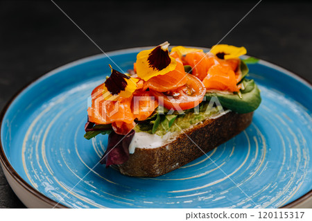 Gourmet smoked salmon toast with fresh vegetables and edible flowers on a vibrant blue plate 120115317