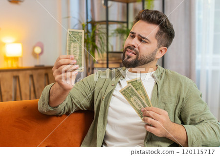 Poor Caucasian young man upset over insufficient amount of money, counting dollar banknotes at home 120115717