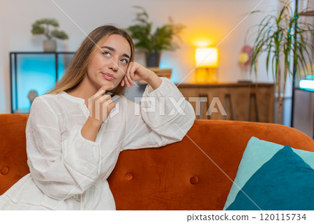 Thoughtful young woman in dress touching chin thinking about where to go for weekend sitting on sofa Thoughtful young woman in dress touching chin thinking about where to go for weekend sitting on sofa 120115734