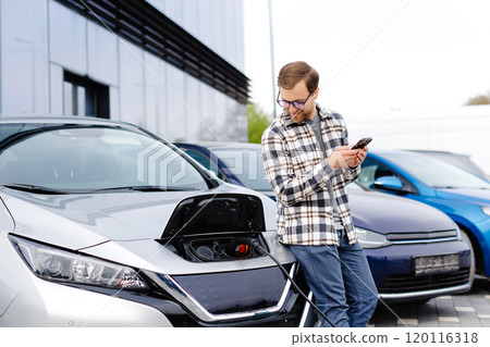 A young man uses a mobile phone while leaning on his electric car while the car is charging 120116318