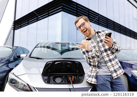 A young man uses a mobile phone while leaning on his electric car while the car is charging A young man uses a mobile phone while leaning on his electric car while the car is charging 120116319
