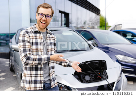A young man stands next to his new electric car and rests while the car is charging 120116321
