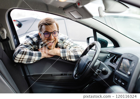 Young man looking through window inside new car, checking it before purchase at modern dealership store. Cheerful guy buying or renting automobile at showroom 120116324