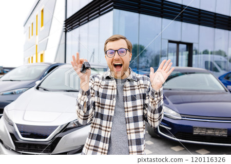 Emotional guy holding key in hand and smiling at camera. Man showing key from his new electric car. Young man buying auto at dealership salon 120116326