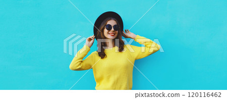 Portrait of happy stylish young woman in black hat, yellow sweater, modern cool girl posing on blue 120116462