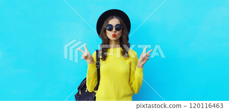 Portrait of stylish young woman in black hat, yellow sweater, modern cool girl posing on blue 120116463