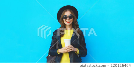 Stylish happy young woman holding cup of coffee in black hat, leather jacket on blue background Stylish happy young woman holding cup of coffee in black hat, leather jacket on blue background 120116464