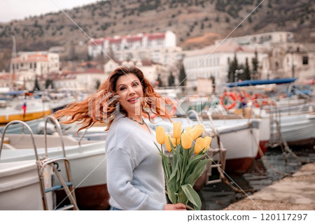 Woman holds yellow tulips in harbor with boats docked in the background., overcast day, yellow sweater, mountains 120117297