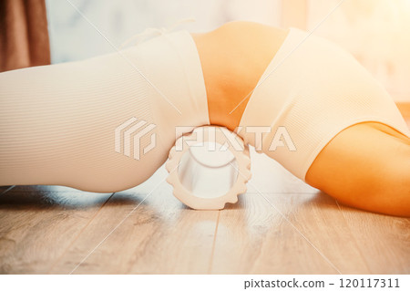 Adult athletic woman, in white bodysuit, performing fascia exercises on the floor - caucasian woman using a massage foam roller - a tool to relieve tension in the back and relieve muscle pain - the 120117311