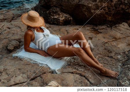Woman Sunbathing Beach Rocks - A woman relaxes on a rocky beach, wearing a white swimsuit and a straw hat, enjoying the sun. Woman Sunbathing Beach Rocks - A woman relaxes on a rocky beach, wearing a white swimsuit and a straw hat, enjoying the sun. 120117318