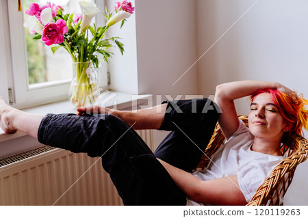 Relaxed young Caucasian woman with colorful hair sitting comfortably in wicker chair. Enjoying quiet moment by window with bouquet of vibrant flowers in vase. Tranquility and peace. 120119263