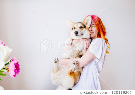 Vibrant young woman with colorful hair embraces her adorable corgi dog on white background. Joyous, loving moment surrounded by flowers, warmth and happiness. Human and pet, love, cheerful living. 120119265
