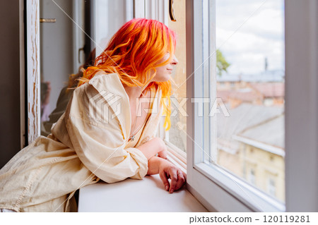 Adult Caucasian woman with colorful red and orange hair looks thoughtfully outside from window, dressed in casual, leans on windowsill, enjoying peaceful moment indoors. 120119281