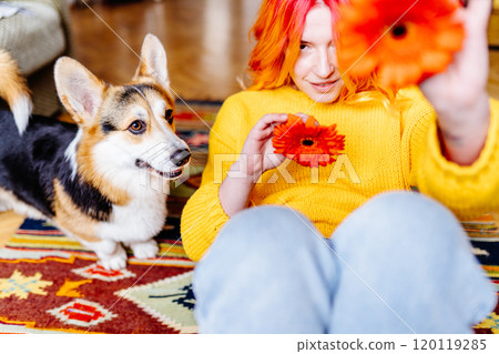 Young Caucasian woman with orange hair holding bright flowers, sitting on floor with playful corgi. Interior setting with colorful rug and cozy environment,joy and companionship. 120119285
