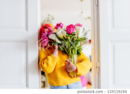 Caucasian woman with vibrant hair holding large bouquet of fresh flowers. Near open door, spring vibes filled with color and life. Home outfit uplifting, joyful atmosphere. 120119288