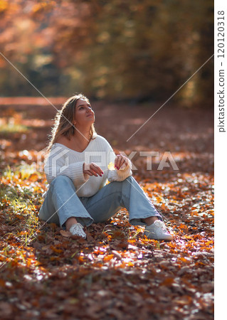 Woman Autumn Leaves Park - A young woman in a white sweater sits amidst fallen leaves in a park during autumn, gazing upwards. 120120318