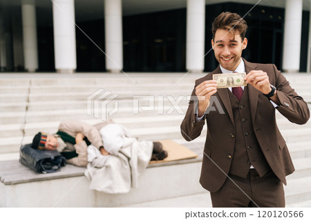Joyful successful businessman in suit cheerful looking at camera as holding dollar bill while homeless person lying on steps of urban building, showing social and economic disparities. Joyful successful businessman in suit cheerful looking at camera as holding dollar bill while homeless person lying on steps of urban building, showing social and economic disparities. 120120566