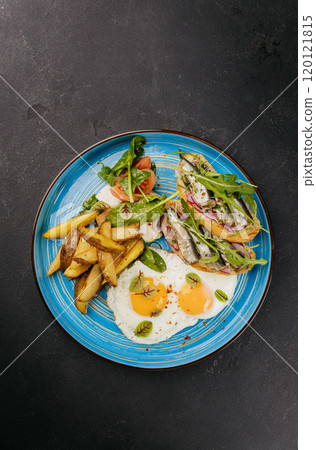 Breakfast plate with fried eggs, roasted potatoes, fresh salad, and sardine toasts on a blue dish Breakfast plate with fried eggs, roasted potatoes, fresh salad, and sardine toasts on a blue dish 120121815