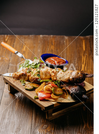 Grilled meat and vegetables on skewers with dipping sauce, served on a rustic wooden board 120121827