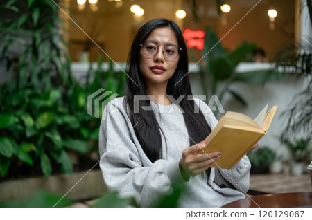 A calm, thoughtful Asian woman reads at an outdoor table in a coffee shop, surrounded by plants. A calm, thoughtful Asian woman reads at an outdoor table in a coffee shop, surrounded by plants. 120129087