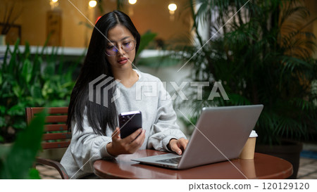 Attractive Asian woman is checking messages on her phone while working remotely from a coffee shop. 120129120