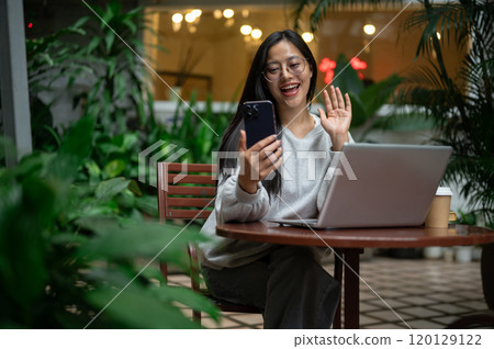 Happy Asian woman is talking on a video call on her phone while working remotely from a coffee shop. 120129122
