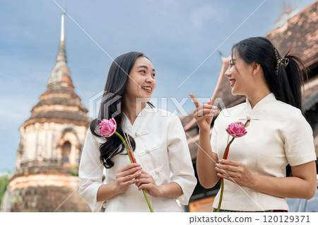 Two beautiful, smiling Asian women stand in a temple, enjoying a conversation together. 120129371