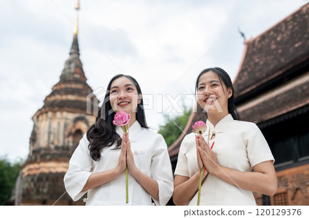 Two beautiful, smiling Asian women stand in a temple, holding pink lotuses in a prayer posture. 120129376