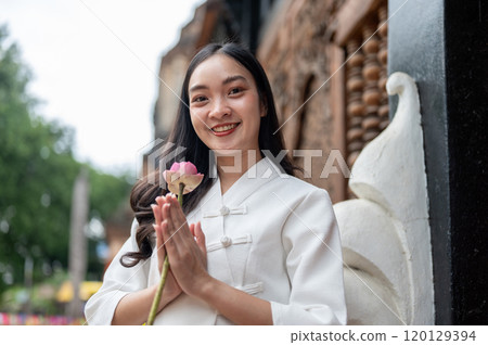 An Asian woman in a traditional dress stands in a temple, holding a lotus in a prayer posture. 120129394