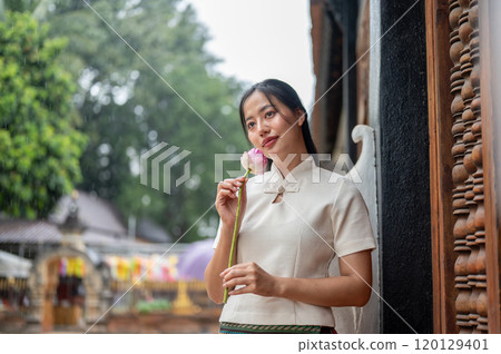 A beautiful Asian woman in a traditional dress stands in a temple, holding a lotus. 120129401