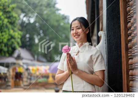 An Asian woman in a traditional dress stands in a temple, holding a lotus in a prayer posture. An Asian woman in a traditional dress stands in a temple, holding a lotus in a prayer posture. 120129402