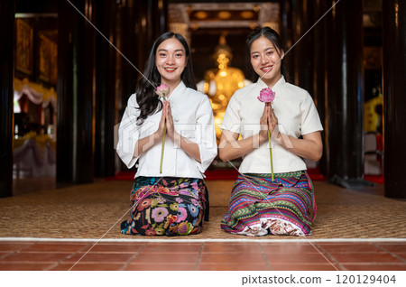 Two charming Asian women in traditional dresses sit in a temple, holding lotuses in a prayer posture 120129404