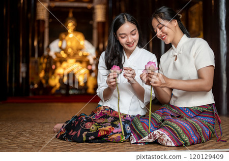 Two beautiful Asian women in traditional dresses sit in a temple, enjoy folding lotus flower petals. 120129409