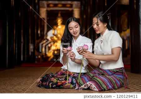 Two beautiful Asian women in traditional dresses sit in a temple, enjoy folding lotus flower petals. Two beautiful Asian women in traditional dresses sit in a temple, enjoy folding lotus flower petals. 120129411