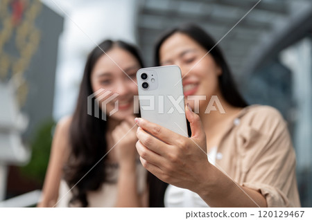 Two happy Asian female friends looking at a smartphone together while standing on a busy street. Two happy Asian female friends looking at a smartphone together while standing on a busy street. 120129467