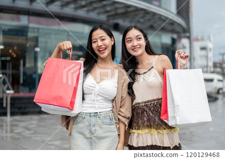 Two Asian women stand in front of the mall, carrying their shopping bags and smiling at the camera. Two Asian women stand in front of the mall, carrying their shopping bags and smiling at the camera. 120129468