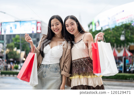 Attractive female friends smile at the camera, carrying shopping bags and standing by a busy street. 120129471