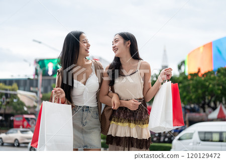 Two attractive Asian female friends smile at each other, carrying their shopping bags by a street. 120129472