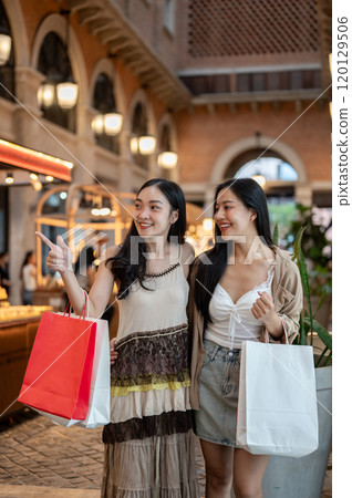 Two charming Asian women enjoying shopping together at a mall, carrying their shopping bags. 120129506