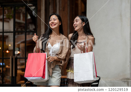 Two happy Asian women walk out of the store with their shopping bags, enjoying shopping together. 120129519