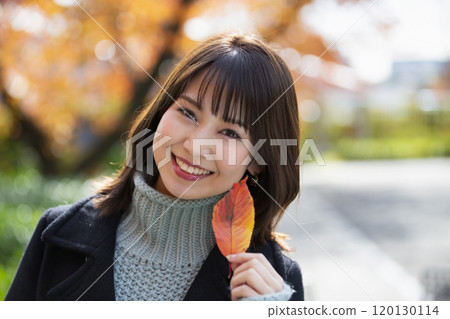A smiling young woman holding a leaf in her left hand with autumn leaves in the background at Sakuranomiya Park in Miyakojima Ward, Osaka City, Osaka Prefecture 120130114
