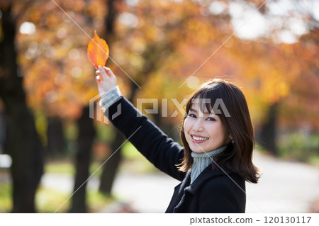 A smiling young woman holding a leaf in her right hand with autumn leaves in the background at Sakuranomiya Park in Miyakojima Ward, Osaka City, Osaka Prefecture 120130117