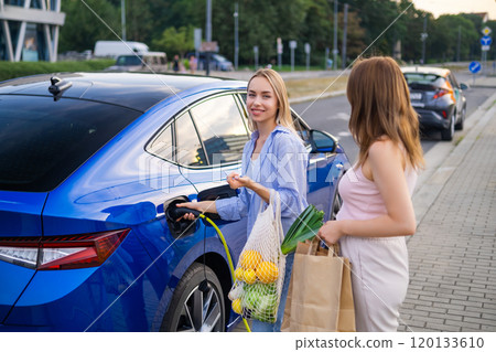Women charging electric car while parked on city street with groceries in the evening. Women charging electric car while parked on city street with groceries in the evening. 120133610