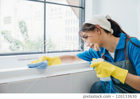 A maid with protective gloves wipes ceramic tiles in a bathroom using a microfiber cloth. Her routine cleaning highlights the essence of purity and hygiene in commercial cleaning. 120133630