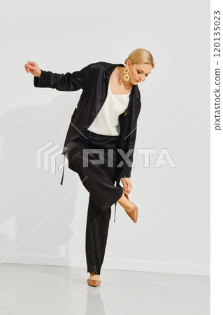 A woman poses elegantly in a stylish black outfit while adjusting her shoe indoors A woman poses elegantly in a stylish black outfit while adjusting her shoe indoors 120135023
