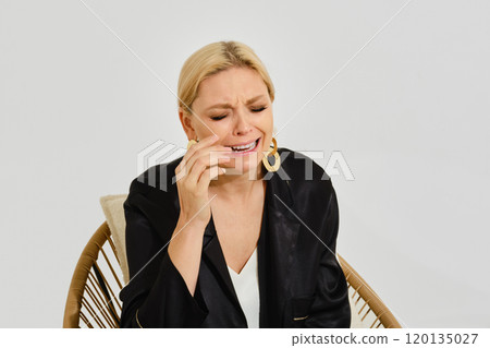 Crying woman expresses feelings while sitting in a modern chair against a plain background Crying woman expresses feelings while sitting in a modern chair against a plain background 120135027