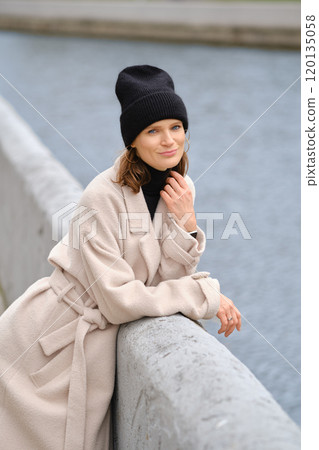 Young woman wearing a black beanie and beige coat poses by a waterway in a modern urban setting Young woman wearing a black beanie and beige coat poses by a waterway in a modern urban setting 120135058