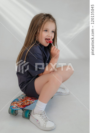a young girl with long blonde hair is sitting on a skateboard, holding a lollipop in her hand. She is wearing a dark T-shirt and sneakers, looking happy and relaxed. a young girl with long blonde hair is sitting on a skateboard, holding a lollipop in her hand. She is wearing a dark T-shirt and sneakers, looking happy and relaxed. 120135845