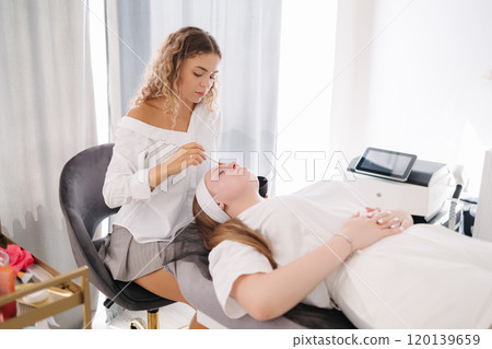 A beautician gently applies a face mask to a woman lying on a spa bed during a facial treatment. Calm atmosphere 120139659